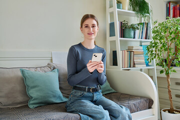 Happy female teenager using smartphone, sitting on couch at home