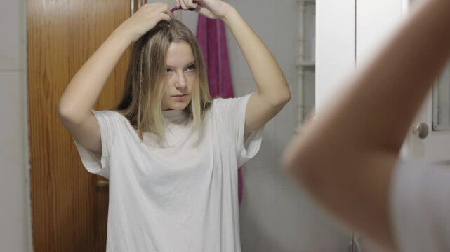 Teenager Girl with long blond hair is looking at her reflection in the bathroom mirror and combing her hair, apparently dissatisfied with how it looks