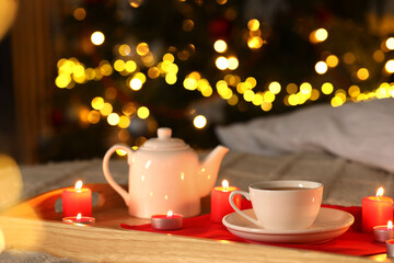 Aromatic tea in cup, teapot and burning candles on bed against blurred Christmas lights, closeup. Bokeh effect
