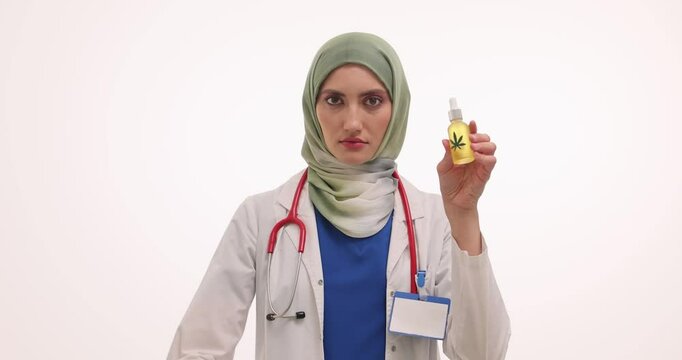Muslim woman doctor shows Stop gesture and CBD oil on white background. Medic in hijab makes denial sign with hand holding marijuana product
