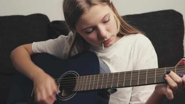 Young Girl sitting on a couch, playing a blue acoustic guitar. Practicing chords and picking patterns, she immerses herself in music, showcasing her passion and dedication