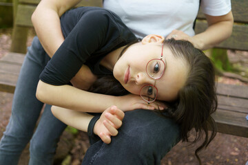 Little sad girl in glasses and T-shirt put her head on her mother's lap in a summer park. Concept...