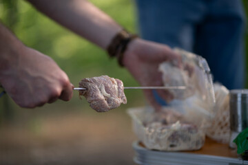 persons hand holds a skewer with raw meat over a barbecue. The background is blurred