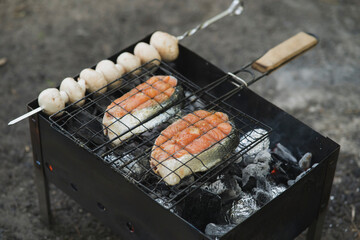 Two salmon steaks and mushrooms on skewers are being grilled over hot coals in a black metal charcoal grill outdoors