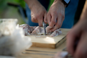 person is preparing raw meat for an outdoor meal. They are using a knife to cut the slices on a wooden cutting board