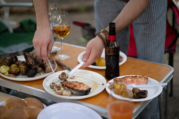 person is serving grilled meat and potatoes on a plate outside. The plate is on a table with other food, including bread and wine