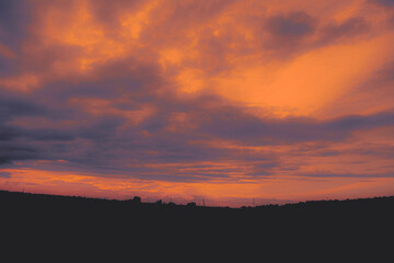 A bright orange sky with beautiful clouds after sunset,  creates a stunning contrast against the dark countryside field. The power lines in the distance add a touch of industrial beauty to the scene.