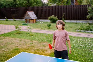 A young boy playing table tennis in a backyard on a sunny day.