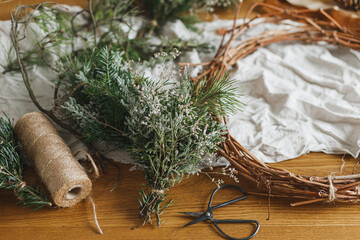 Making Christmas rustic wreath. Evergreen branches, pine cones, twigs, twine, scissors on rustic table in atmospheric festive room. Winter holiday preparations