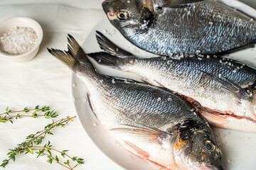 Raw fresh sea bream fish with salt and oregano on a white plate and a light background, ready to cook.