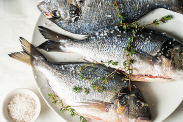 Raw fresh sea bream fish with salt and oregano on a white plate and a light background, ready to cook.