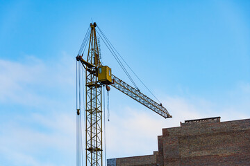 A tall yellow crane stands prominently on a construction site, surrounded by an industrial structure. It is actively lifting materials against a bright blue sky