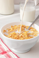 Filling a white bowl with milk with cereals on a withe kitchen table.