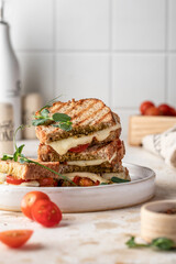 Grilled sandwich panini with mozzarella cheese, cherry tomatoes and pesto on white plate, kitchen table background. Delicious breakfast or snack, traditional italian food