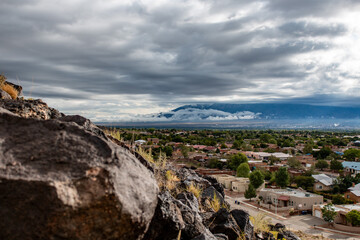 A view of Albuquerque from Petroglyph National Monument, featuring storm clouds and a rocky foreground.
