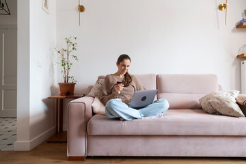 Woman sitting on sofa is shopping online using laptop and credit card