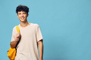 Young man smiling against a light blue background, wearing a casual beige t shirt and carrying a yellow backpack, embodying a sense of youth and adventure in urban style