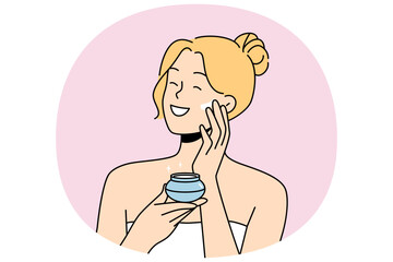 Woman applies anti-aging cream to face to stay beautiful, standing in bath towel after shower. Happy blonde girl smiles and uses cosmetic cream to help prevent wrinkles and pimples.