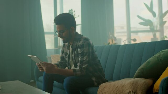 Happy young man with glasses use tablet computer sitting on sofa at home smiling. Thinking. Communication. Camera moving around. 360 degrees