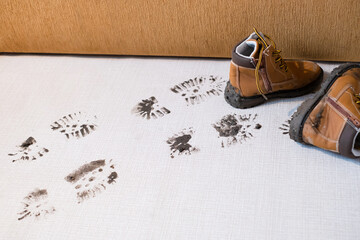 Muddy footprints trail across a clean floor, with a pair of brown boots resting nearby. Dirty traces of shoes on the carpet or a rug near the sofa. Space for text