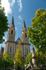 A majestic church with twin spires rises against a bright blue sky, surrounded by lush green trees and a decorative fountain featuring classical statues in the foreground.