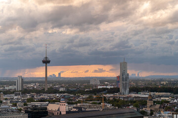 A cityscape at sunset with dramatic clouds, a prominent TV tower, modern skyscrapers, and a sprawling urban environment. The horizon features wind turbines and an industrial skyline.