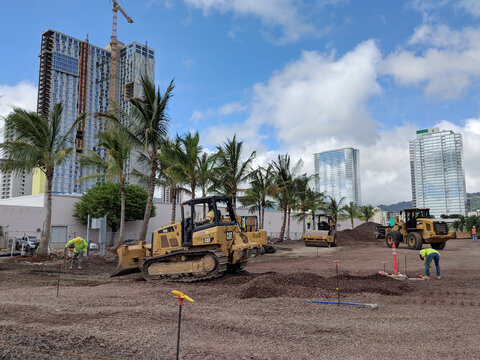 Construction work underway at a site in Honolulu with heavy machinery and workers amid tall buildings and palm trees