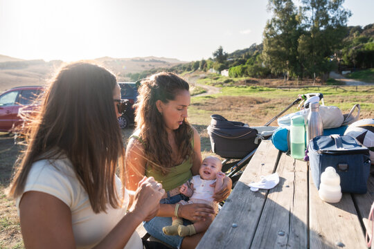 Mother holding smiling baby while talking to friend at picnic table