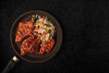 fried chicken fillet steaks with rice with vegetables on metal frying pan on black textured background. artistic dark food photo with copy space top view