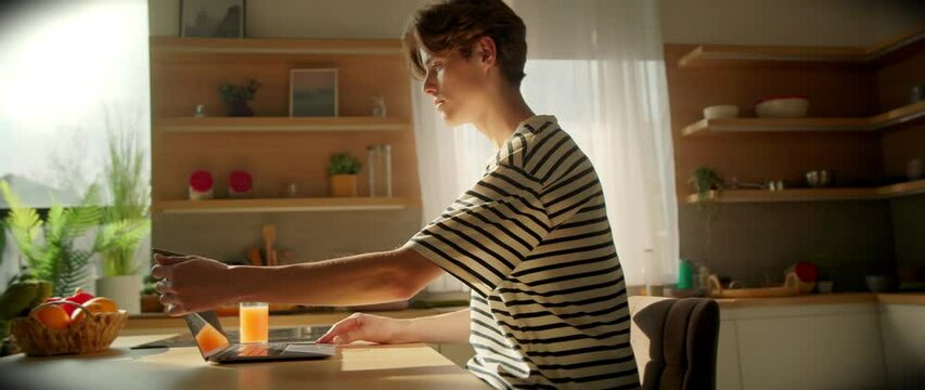 Tracking shot of young male student sitting at table opening laptop and typing on keyboard in kitchen. Smiling guy waving to camera via video call. Education concept. Distance learning. Indoors