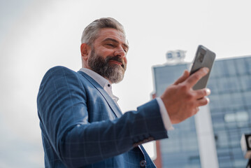 A happy mature business man using a mobile phone outside with urban buildings in the background