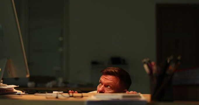 Frightened man hides under desk glancing around in dark office. Employee feels vulnerable and loneliness staying late at stormy weather