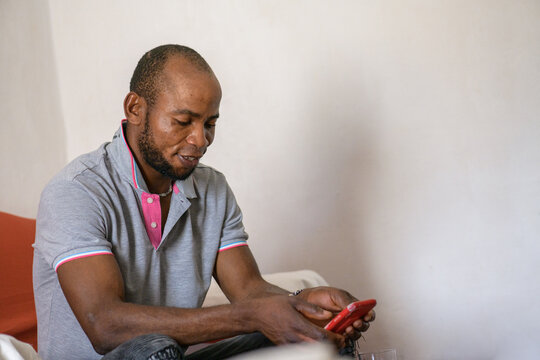 Positive nigerian man using smartphone and sitting on red sofa