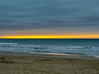 Golden Sunrise Over the Mediterranean Sea at Palavas-les-Flots, France, with a Small Sailboat on the Horizon, Gentle Waves, Sandy Beach, and Dramatic Overcast Clouds