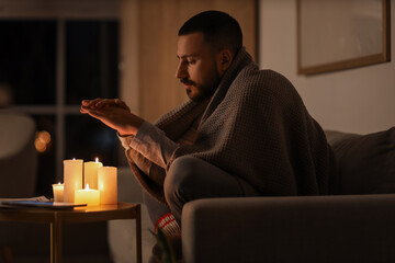 Frozen young man with plaid warming hands near burning candles at home during blackout