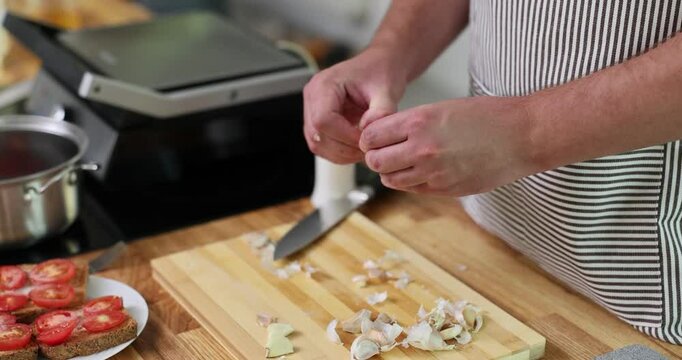 Man peels garlic cloves standing near wooden countertop. Male chef prepares ingredients cooking delicious meal in domestic kitchen slow motion