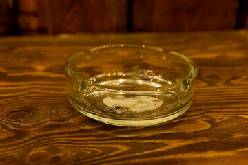 Clear glass ashtray sits on a rustic wooden table with remnants of tobacco at a casual gathering spot in the evening