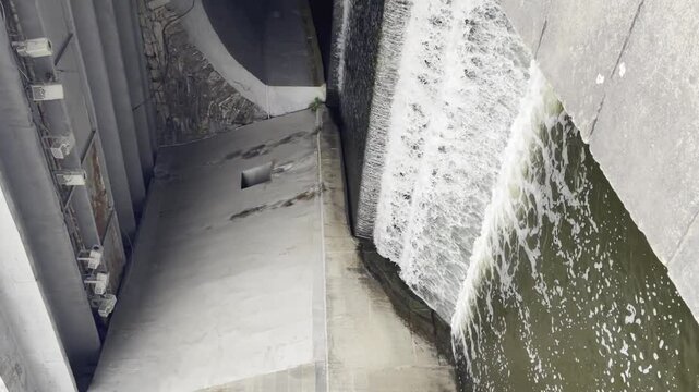 Vertical video small grey concrete dam in city with waterfall moving into river