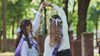 Two students in graduation sashes joyfully dance together in the schoolyard to celebrate their last day of school