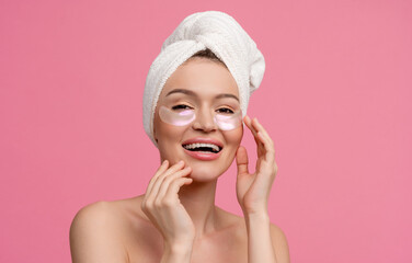 smiling h woman after shower on pink background with bath towel doing morning skincare routine using eye patches