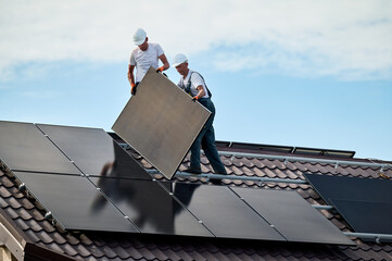 Workers building solar panel system on rooftop of house. Two men installers in helmets installing...