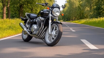 Close-up view of a black motorcycle's front wheel and headlight on a quiet asphalt road surrounded by lush green trees in soft light