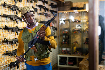 African-american man wearing military vest and holding machine gun in air weapon shop.