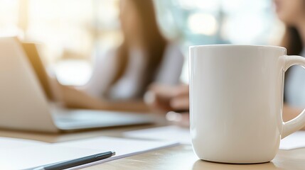 A focus on a white coffee mug in a collaborative workspace with two women blurred in the background.