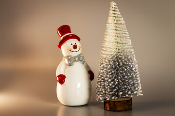 Festive New Year decoration, decor of a ceramic snowman with a red hat and a bow on a gray background with a cone-shaped Christmas tree with sparkles.