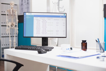 Empty doctors office with healthcare equipment and cabinet neatly arranged for use. Computer displaying medical data is on a desk in clinical room, along with a clipboard, thermometer and pill bottle.