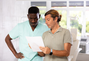Before cosmetology manipulation, man patient sits in chair and reads documents with male doctor...