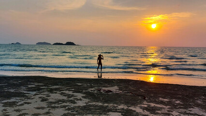 Woman standing in shallow ocean water, admiring vibrant sunset over calm sea with distant islands