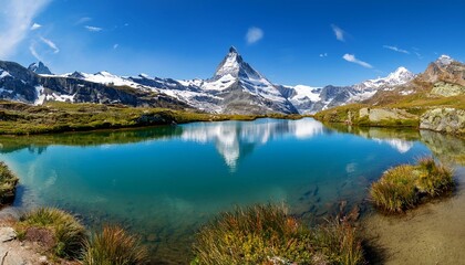 Naklejka premium Panorama of the Alps: Clear mountain lake with Matterhorn in the background, surrounded by lush greenery and snow-capped peaks under a blue sky