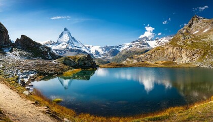 Naklejka premium Panorama of the Alps: Clear mountain lake with Matterhorn in the background, surrounded by lush greenery and snow-capped peaks under a blue sky